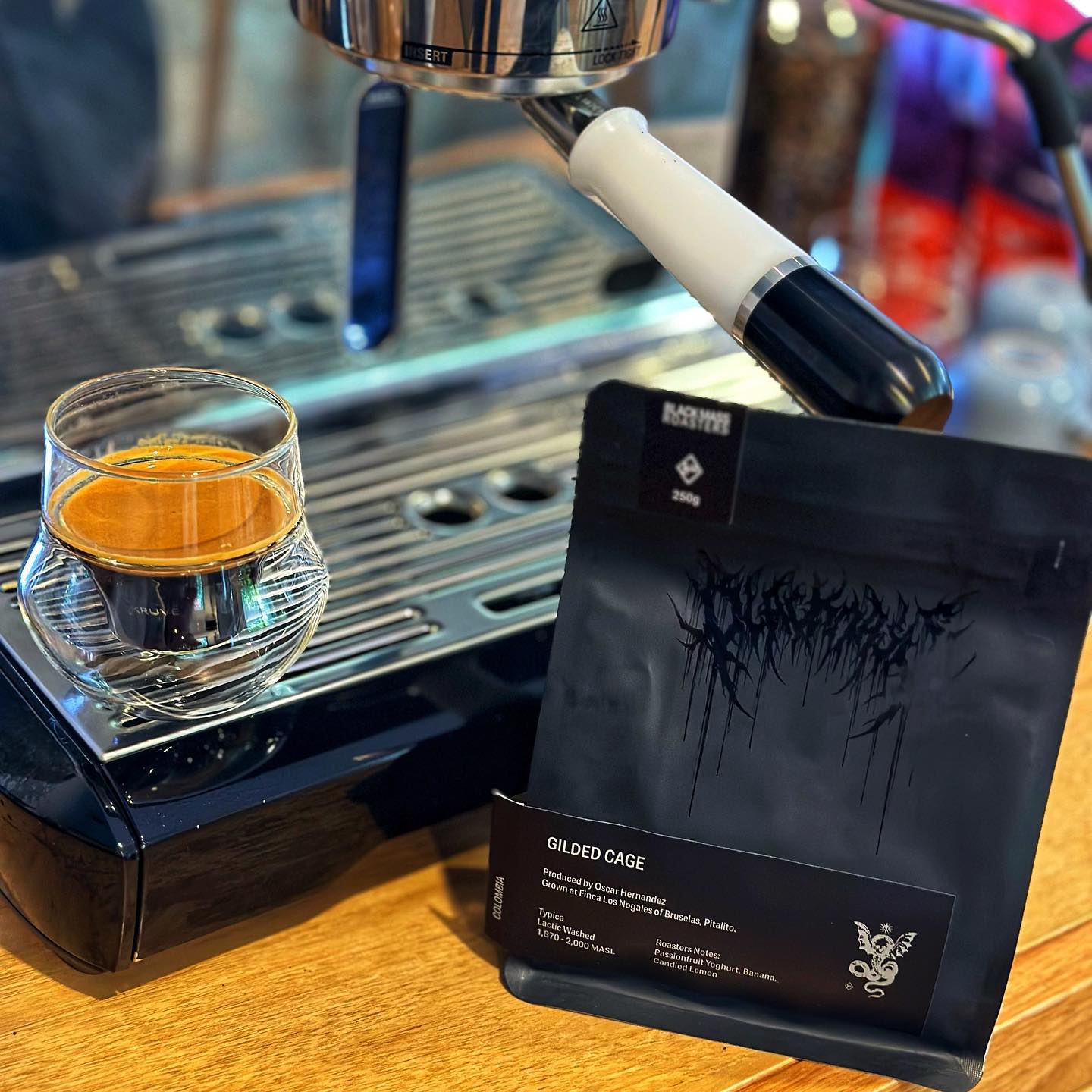 An espresso in a glass sitting on a coffee machine next to a bag of the Black Mass beans. The bag is black with a metal band style logo embossing