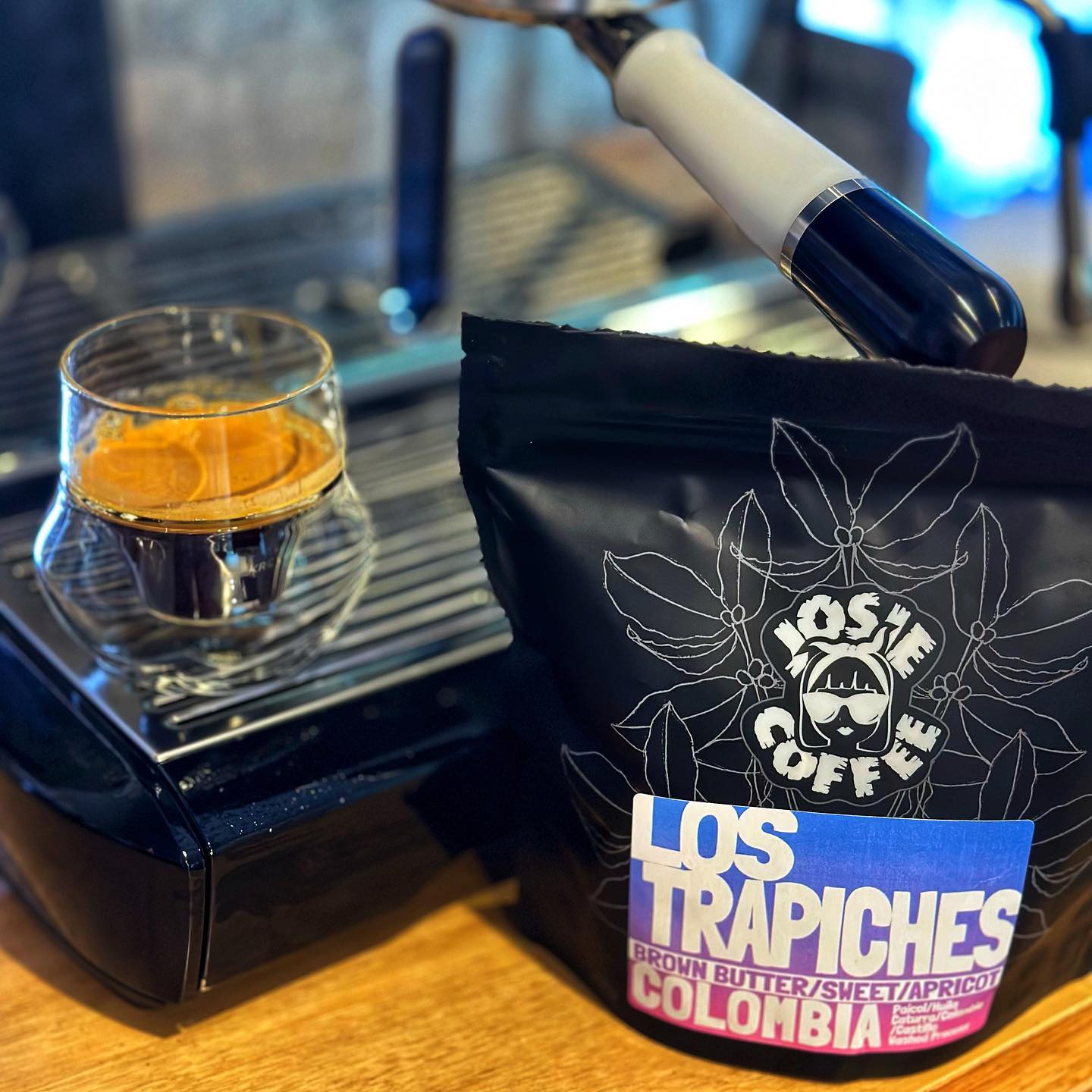 An espresso in a glass sitting on a coffee machine next to a bag of the Josie Coffee Los Trapiches beans.