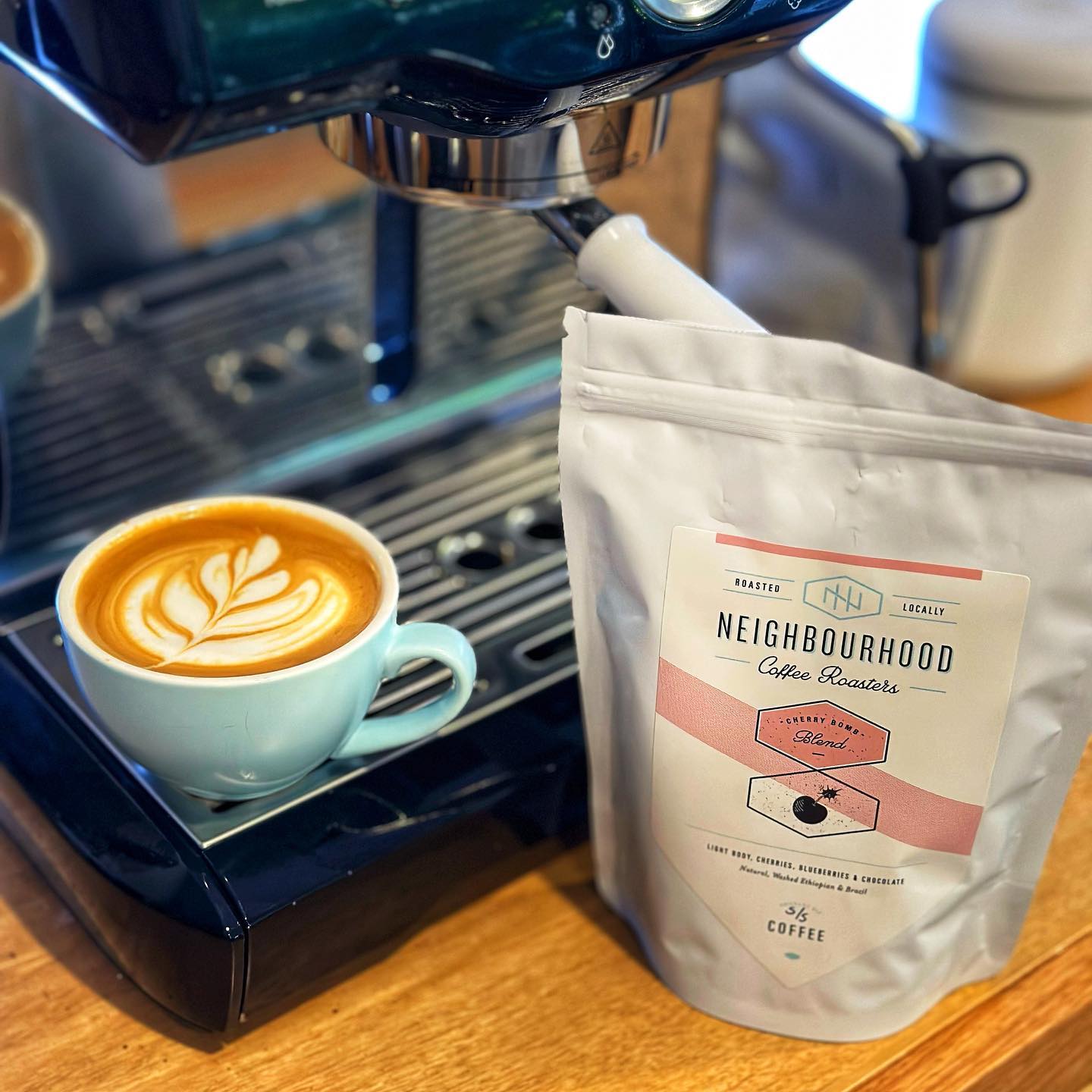 A flat white coffee with latte art sitting on a coffee machine next to a bag of the Neighbourhood Coffee Roasters Cherry Bomb beans