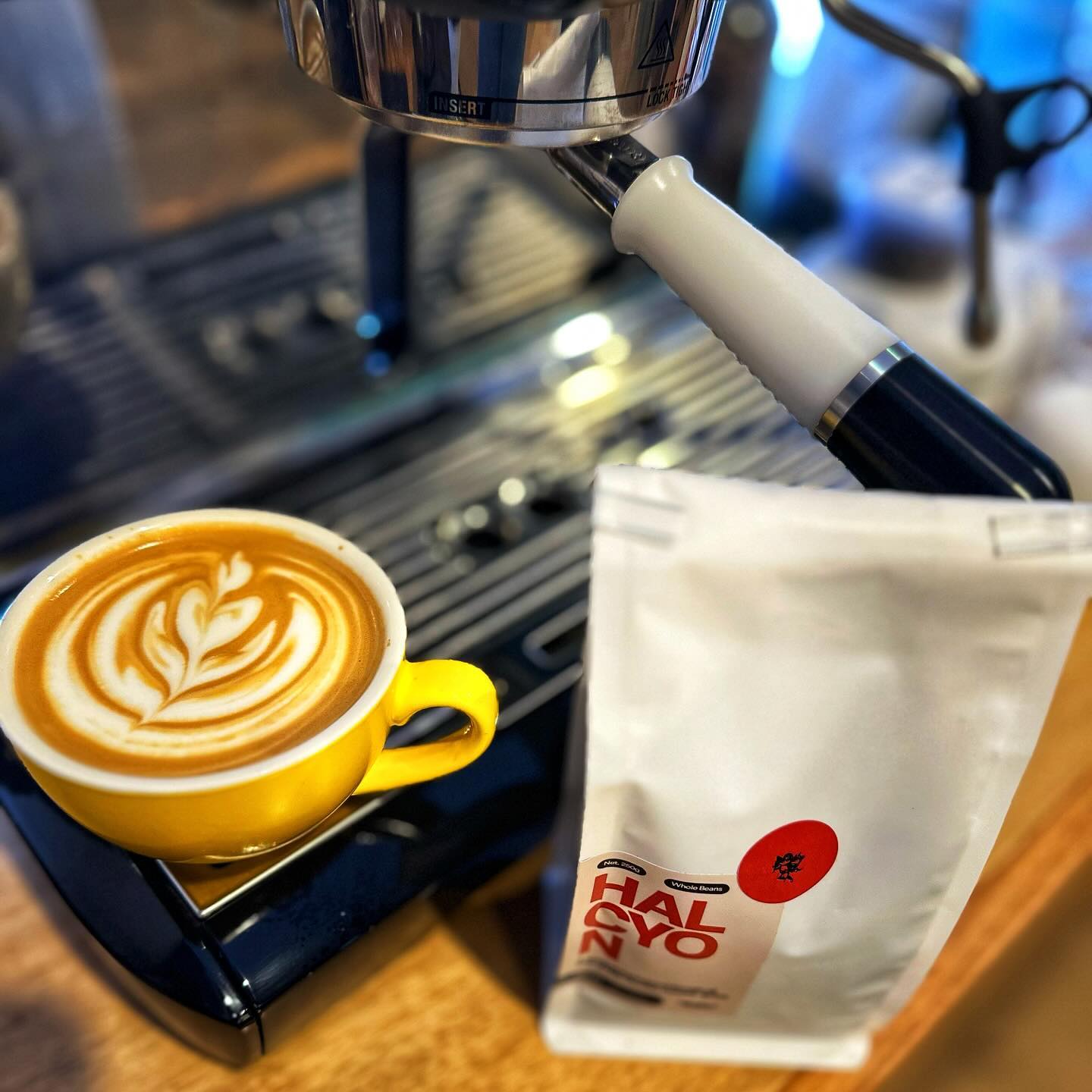 A flat white coffee with latte art sitting on a coffee machine next to a bag of the Omen Coffee Supply Halcyon beans