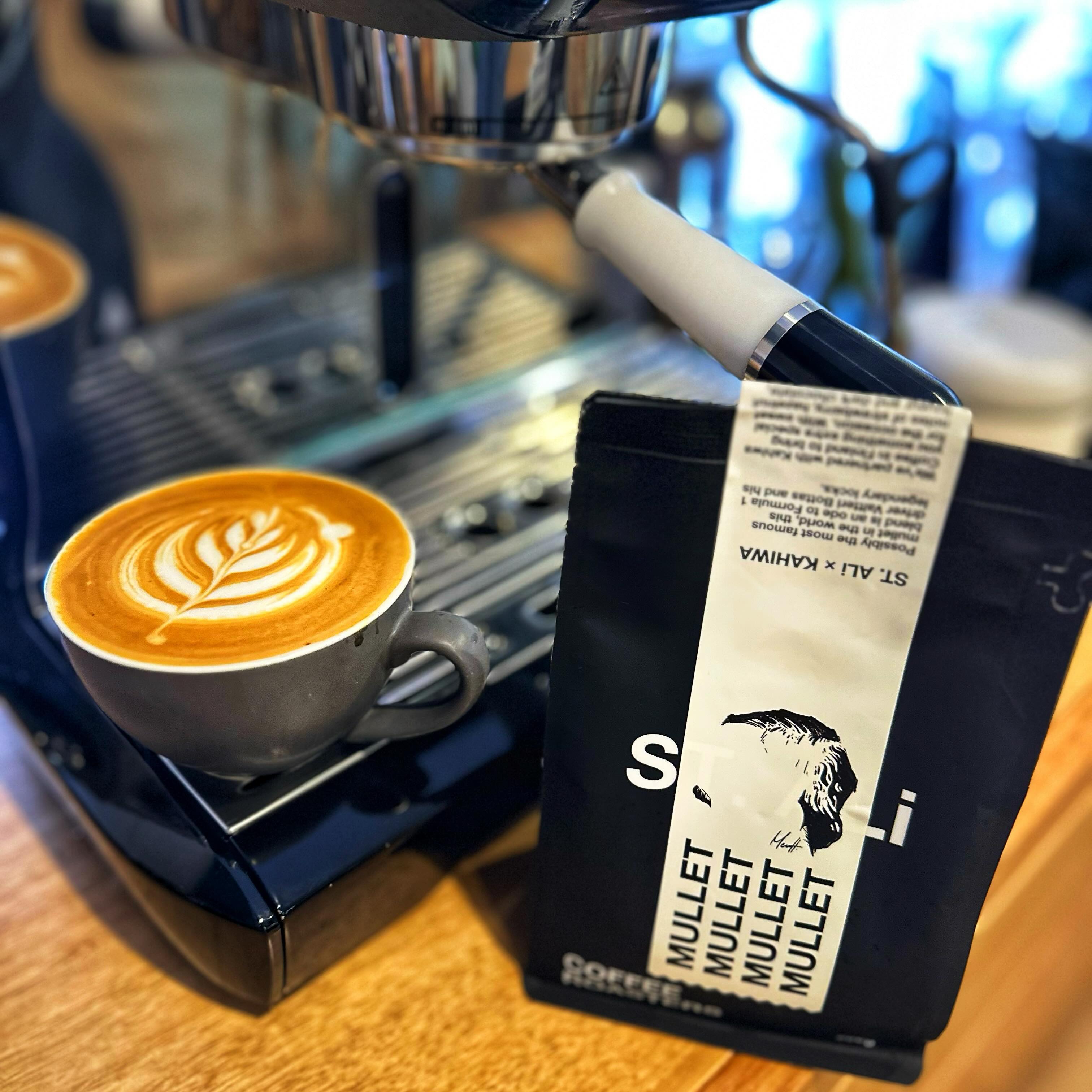 A flat white coffee with latte art sitting on a coffee machine next to a bag of the Mullet coffee beans