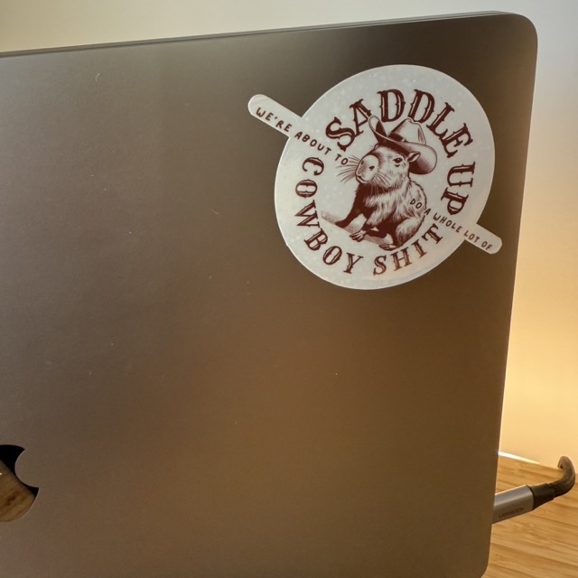 A Macbook with a sticker on it. The sticker is a Capybara in a cowboy hat and the lettering says 'Saddle up, we're about to do a whole lot of cowboy shit